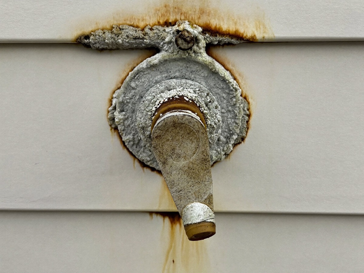 A badly corroded outdoor hose bib crusted with salt deposits, rust streaking down the siding beneath it
