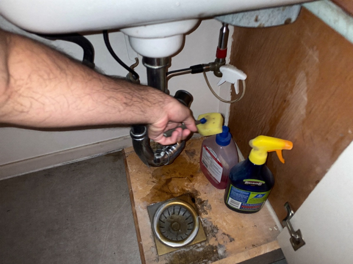 A plumber's hands working a wrench on a corroded drain trap under a kitchen sink cabinet